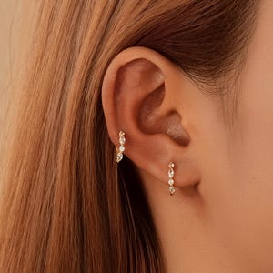 May include: Close-up of two gold hoop earrings, each adorned with clear, sparkling gemstones. One earring is in the upper ear, the other in the lower ear. The earrings are set against the backdrop of a person's ear and hair.