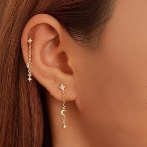 Puede incluir: Pendientes celestiales dorados con estrellas, lunas crecientes y detalles de diamantes. Un pendiente es un perno con una cadena colgante, mientras que el otro es una cadena que cuelga de la oreja. Los pendientes se usan en la oreja de una persona.