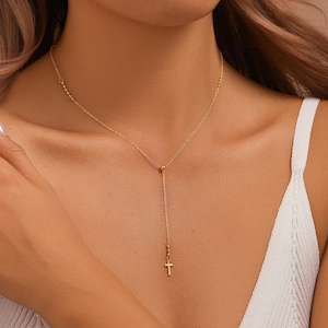 Op de afbeelding: Een delicate goudkleurige ketting met een kruisvormige hanger. De ketting heeft een Y-vormig ontwerp, met een klein kruis aan het einde van de verticale drop. De ketting heeft kleine kralenaccenten aan de bovenkant.