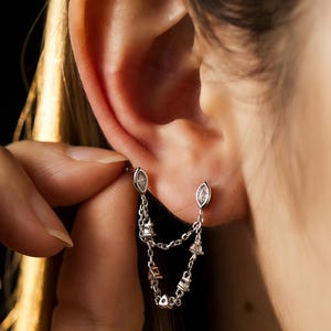 May include: Silver double-chain earrings with clear, oval-shaped gems at the top and dangling chains. The chains connect to various small, geometric gems. The earrings are shown on an ear against a dark background.