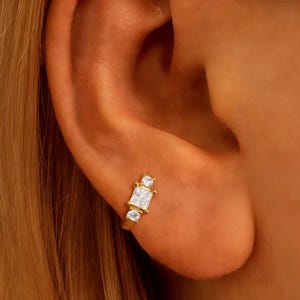 Peut inclure: Boucle d'oreille dorée ornée d'une pierre rectangulaire transparente au centre, flanquée de deux petites pierres rondes et transparentes. La boucle d'oreille est portée sur une oreille.