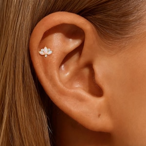 Könnte beinhalten: Nahaufnahme eines Ohrs mit einem goldfarbenen Ohrstecker. Der Ohrring hat einen Cluster aus drei opalartigen Steinen und zwei kleinen klaren Steinen. Der Ohrring ist in einem goldfarbenen Metall gefasst.