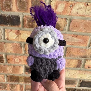 May include: A crocheted plush figure featuring a light purple body, a dark gray lower section, and black feet. It has a single large central eye with a white and gray border, black side bands, and a tuft of vibrant purple yarn hair on top.