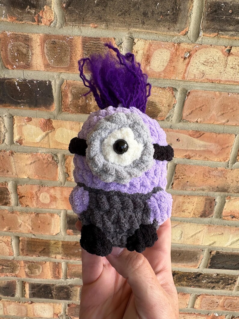 May include: A crocheted plush figure featuring a light purple body, a dark gray lower section, and black feet. It has a single large central eye with a white and gray border, black side bands, and a tuft of vibrant purple yarn hair on top.