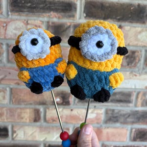 May include: Two crocheted plush figures resembling yellow characters with blue overalls, black feet, and a single large eye with a grey frame. Each figure is attached to a thin metal rod featuring small red and blue beads at the bottom handle.