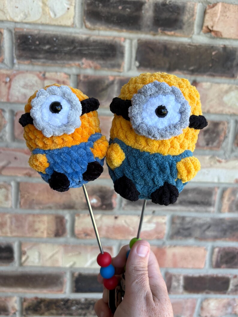 May include: Two crocheted plush figures resembling yellow characters with blue overalls, black feet, and a single large eye with a grey frame. Each figure is attached to a thin metal rod featuring small red and blue beads at the bottom handle.