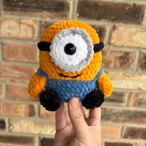 Puede incluir: Un juguete de peluche hecho a mano de un personaje Minion, con un cuerpo amarillo, un peto azul y zapatos negros. El Minion tiene un ojo grande y &uacute;nico con un marco blanco y gris y gafas negras. El juguete est&aacute; hecho de hilo suave y texturizado.