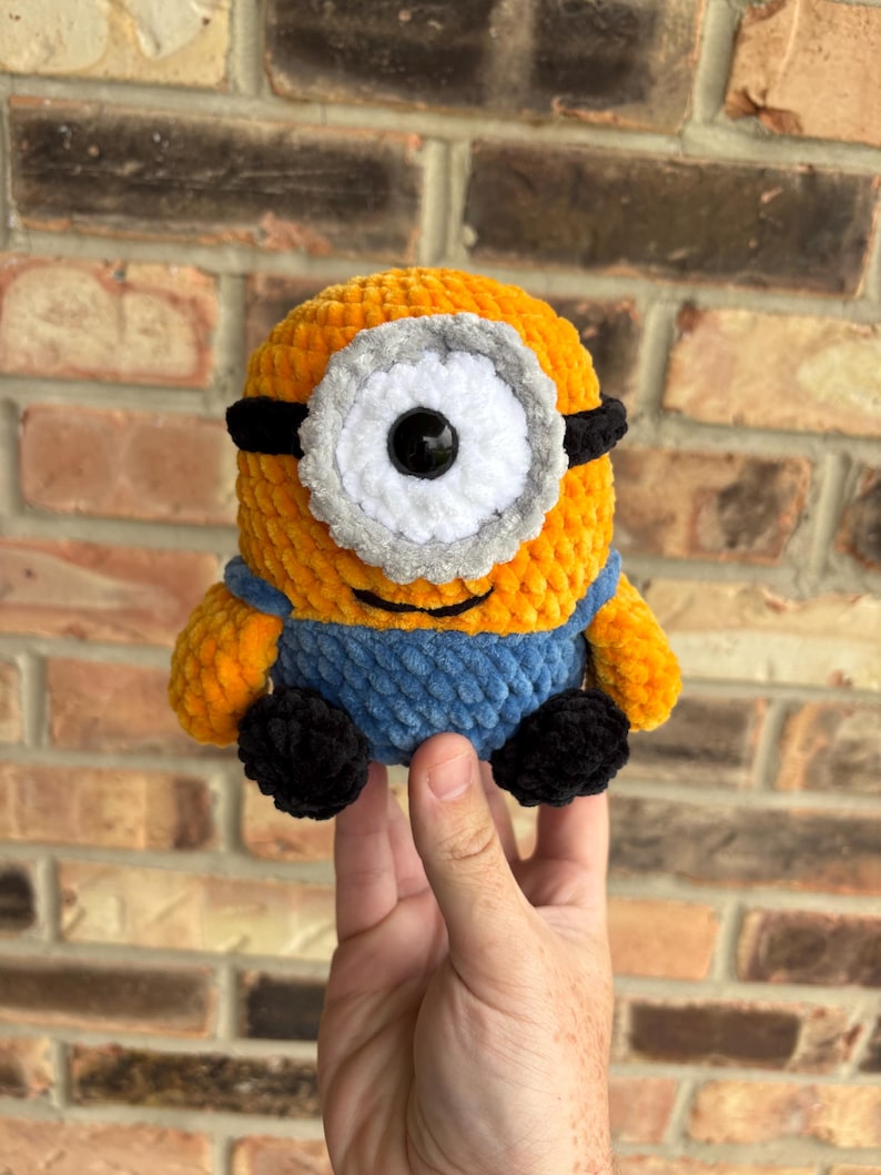 Puede incluir: Un juguete de peluche hecho a mano de un personaje Minion, con un cuerpo amarillo, un peto azul y zapatos negros. El Minion tiene un ojo grande y &uacute;nico con un marco blanco y gris y gafas negras. El juguete est&aacute; hecho de hilo suave y texturizado.