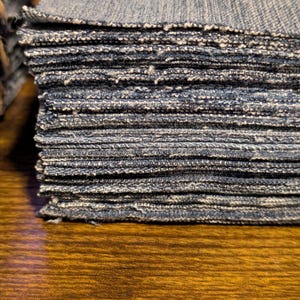 May include: A stack of square denim fabric pieces, showing the raw edges and the texture of the blue jean material. The fabric is stacked on a wooden surface.