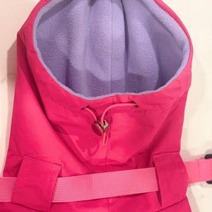 May include: A bright pink hooded garment featuring a soft light purple fleece lining. The hood includes a pink drawstring with a metallic toggle fastener for adjustment. A matching pink fabric strap with a buckle is visible across the front.