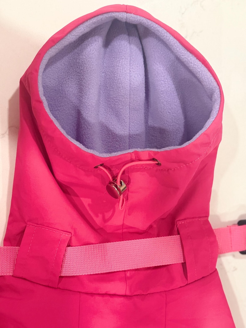 May include: A bright pink hooded garment featuring a soft light purple fleece lining. The hood includes a pink drawstring with a metallic toggle fastener for adjustment. A matching pink fabric strap with a buckle is visible across the front.