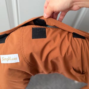 May include: A rust-colored garment featuring a white rectangular label with the text SimpliLuxe printed in gold. The fabric includes black hook-and-loop fastener strips along the top edge, designed for securing the item in place.
