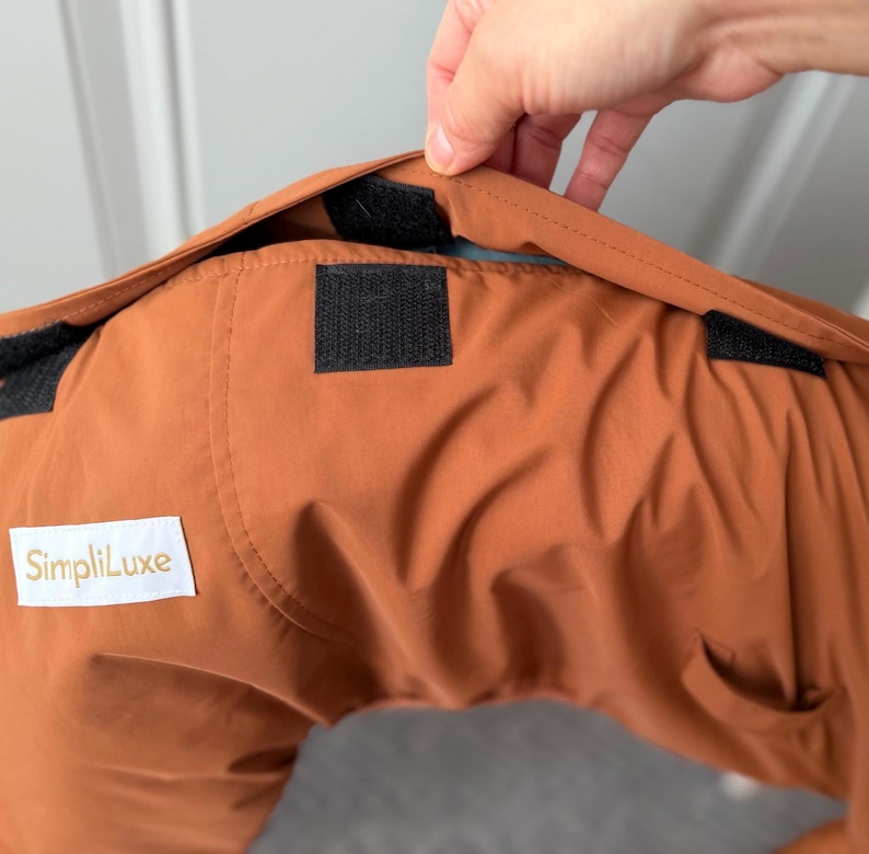 May include: A rust-colored garment featuring a white rectangular label with the text SimpliLuxe printed in gold. The fabric includes black hook-and-loop fastener strips along the top edge, designed for securing the item in place.