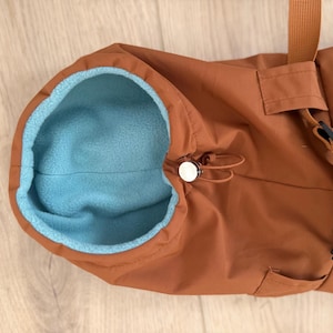May include: A rust-colored outer garment featuring a bright light blue fleece lining. The piece includes a drawstring closure with a silver-toned toggle at the neck opening and a matching rust-colored strap attached to the side of the main body.