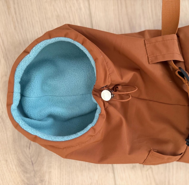 May include: A rust-colored outer garment featuring a bright light blue fleece lining. The piece includes a drawstring closure with a silver-toned toggle at the neck opening and a matching rust-colored strap attached to the side of the main body.