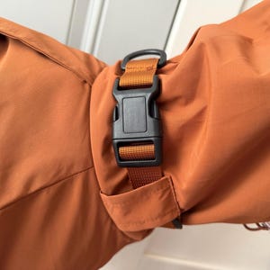 May include: A rust-colored fabric garment featuring a black plastic side-release buckle attached to a matching rust-colored nylon webbing strap. The strap is integrated into the sleeve or cuff area of the garment, providing an adjustable closure.