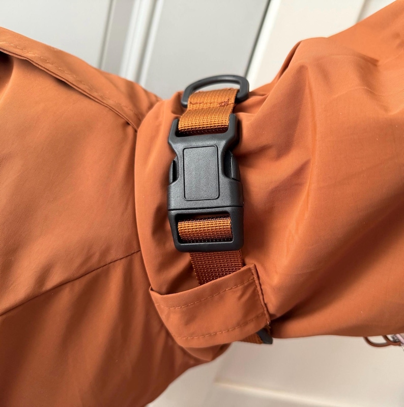 May include: A rust-colored fabric garment featuring a black plastic side-release buckle attached to a matching rust-colored nylon webbing strap. The strap is integrated into the sleeve or cuff area of the garment, providing an adjustable closure.
