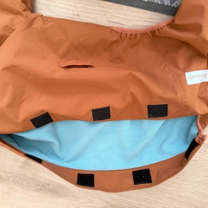 May include: A rust-colored fabric cover featuring a light blue fleece lining, multiple black hook-and-loop fastener strips along the edges, and a white rectangular label with the text Spillluxe printed in black on the upper right side.