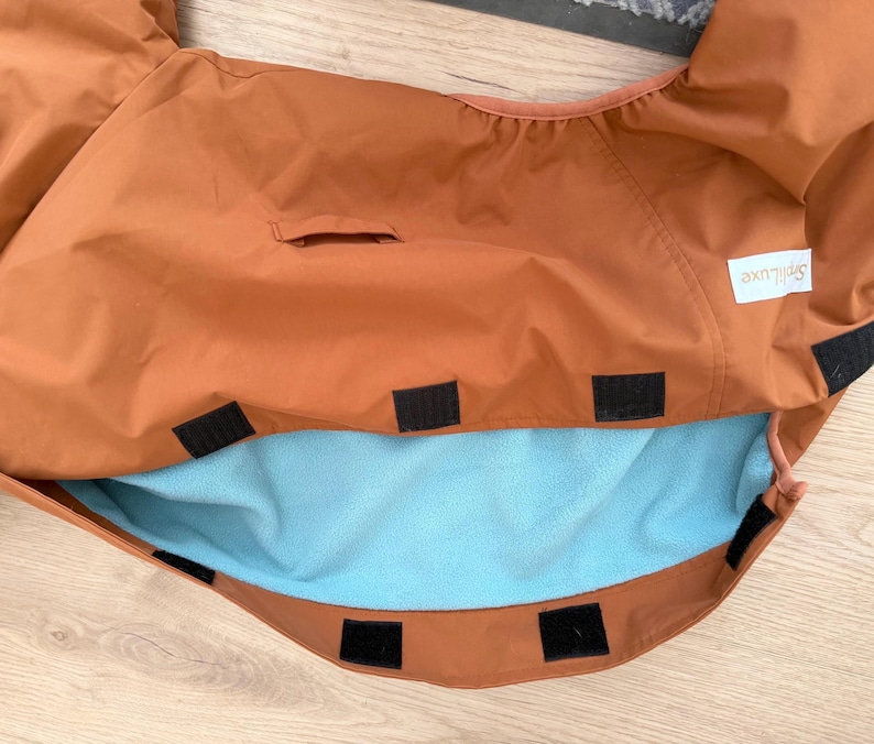 May include: A rust-colored fabric cover featuring a light blue fleece lining, multiple black hook-and-loop fastener strips along the edges, and a white rectangular label with the text Spillluxe printed in black on the upper right side.