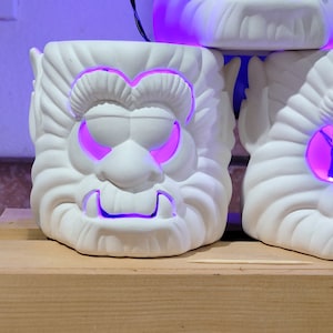 May include: Three white ceramic candle holders shaped like cartoon monster faces with purple light shining through the eye and mouth cutouts.