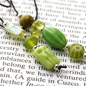 May include: A handmade necklace featuring a variety of green glass beads in different shapes and sizes, strung on a black cord. The beads include round, oval, and textured designs, with silver-toned accents. The necklace is displayed on a printed page.