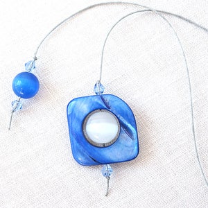 May include: A blue and white square-shaped bookmark with a silver ring and a white circle in the center. The bookmark is attached to a silver cord with blue crystal beads.