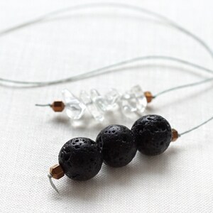 Może przedstawiać: Naszyjnik z trzema czarnymi koralikami z lawy wulkanicznej i sznurem przezroczystych kryształów kwarcu. Naszyjnik wykonany jest z cienkiego szarego sznurka i małych miedzianych koralików.