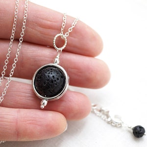 Może przedstawiać: Srebrny naszyjnik z okrągłą zawieszką z czarnego kamienia lawowego. Zawieszka jest osadzona w srebrze i zawieszona na delikatnym srebrnym łańcuszku. Naszyjnik jest prezentowany w dłoni, ukazując design i łańcuszek.