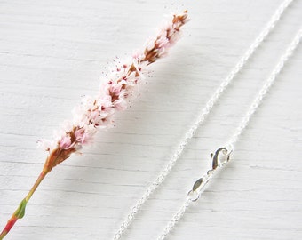 May include: A silver chain necklace with a lobster clasp closure. The chain is on a white wooden surface with a sprig of pink flowers.