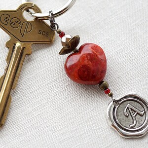 May include: A red heart-shaped charm with a silver wax seal charm with the letter 'A' on it. The charms are attached to a key ring with a brass key.
