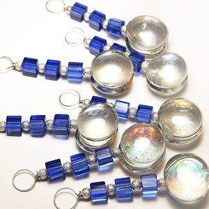 May include: Several decorative charms with a silver loop, clear glass orb, and blue square beads. The beads are strung together with silver-colored spacers. The orbs have a reflective, iridescent quality.