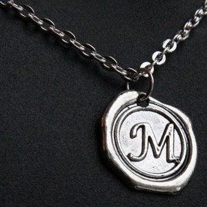 May include: Silver-toned necklace featuring a pendant with the letter "M" in a circular design. The pendant has a textured, antique appearance, and is suspended from a delicate chain. The background is a dark, solid color.
