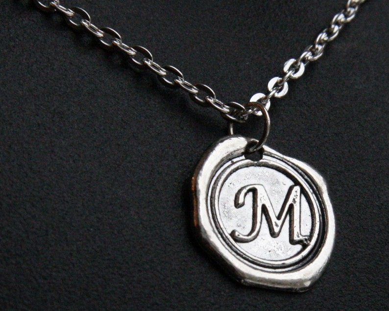 May include: Silver-toned necklace featuring a pendant with the letter "M" in a circular design. The pendant has a textured, antique appearance, and is suspended from a delicate chain. The background is a dark, solid color.