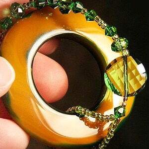May include: A gold-toned ring with a green and gold beaded design. The ring is made of a smooth, polished material and has a large, faceted green gemstone.