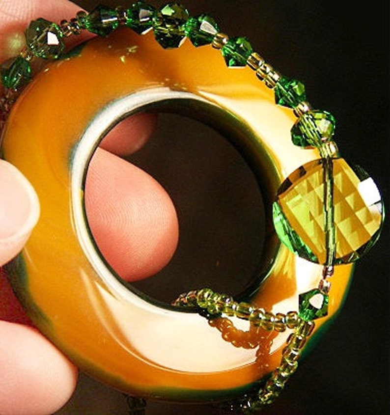 May include: A gold-toned ring with a green and gold beaded design. The ring is made of a smooth, polished material and has a large, faceted green gemstone.