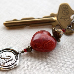May include: A red heart-shaped charm with a silver charm with the letter A on it, attached to a gold key with a key ring. The key ring has a red and silver bead.