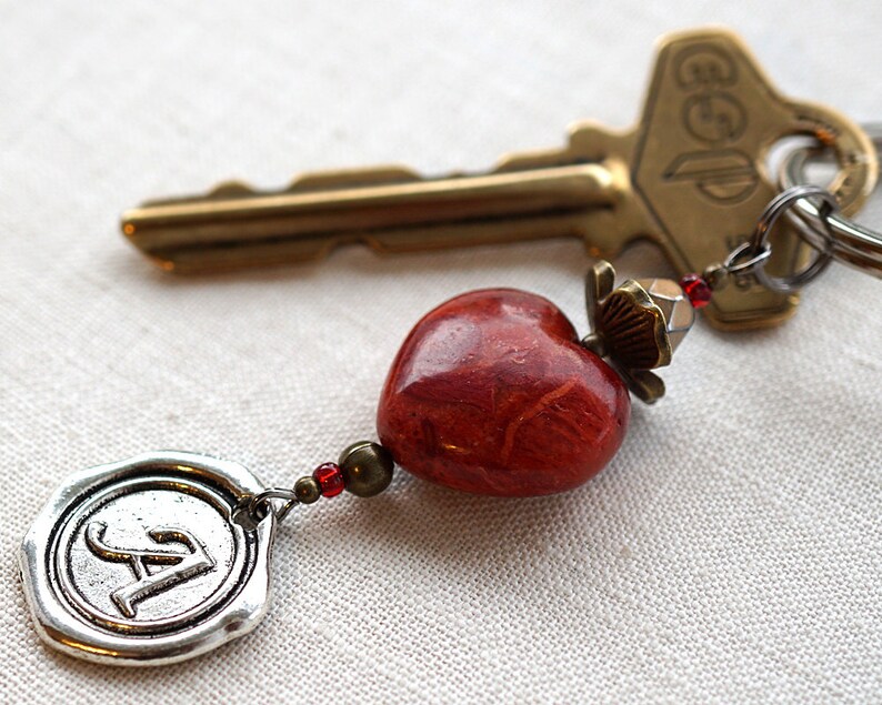 May include: A red heart-shaped charm with a silver charm with the letter A on it, attached to a gold key with a key ring. The key ring has a red and silver bead.