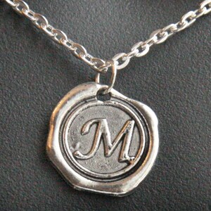 May include: A silver-toned necklace featuring a pendant with the letter "M" in a decorative script. The pendant is circular, resembling a wax seal, and is attached to a delicate chain. The background is a dark, textured surface.
