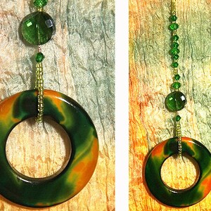 May include: A green and orange marbled ring pendant hangs from a beaded chain with green glass beads. The pendant is suspended against a textured background of orange, yellow, and green.