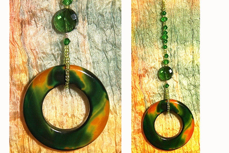 May include: A green and orange marbled ring pendant hangs from a beaded chain with green glass beads. The pendant is suspended against a textured background of orange, yellow, and green.
