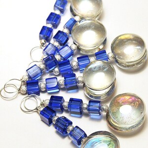 May include: Close-up of several handmade jewelry pieces. Each piece features a combination of blue cube-shaped beads, silver-colored spacer beads, and clear, iridescent glass beads. The pieces are connected with silver-colored wire loops.