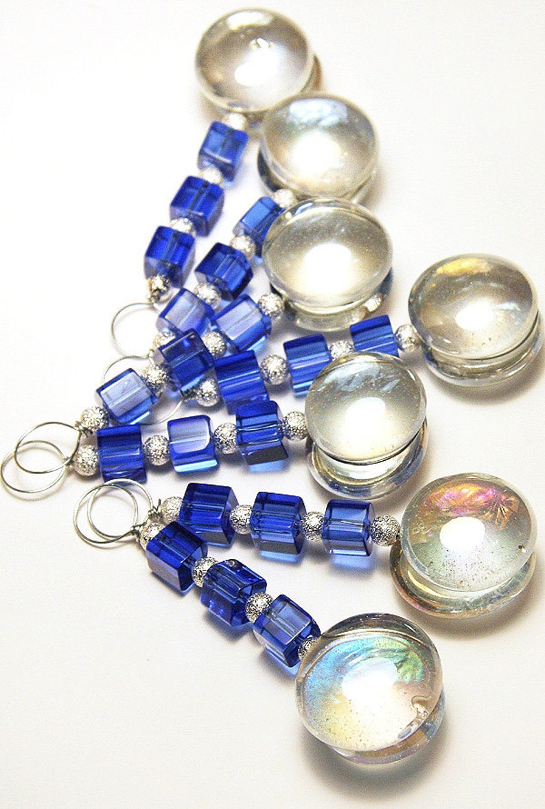 May include: Close-up of several handmade jewelry pieces. Each piece features a combination of blue cube-shaped beads, silver-colored spacer beads, and clear, iridescent glass beads. The pieces are connected with silver-colored wire loops.
