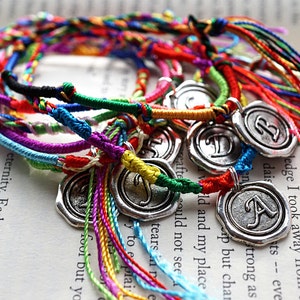 Peut inclure: Gros plan sur un groupe de bracelets d'amitié colorés avec des breloques argentées. Chaque breloque porte la lettre 'A'. Les bracelets sont faits de cordons fins et tressés de différentes couleurs, notamment rouge, bleu, vert, jaune et violet.
