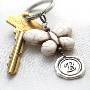 May include: A white butterfly charm with a brown and silver key chain. The charm has a silver seal with the letter "B" on it.