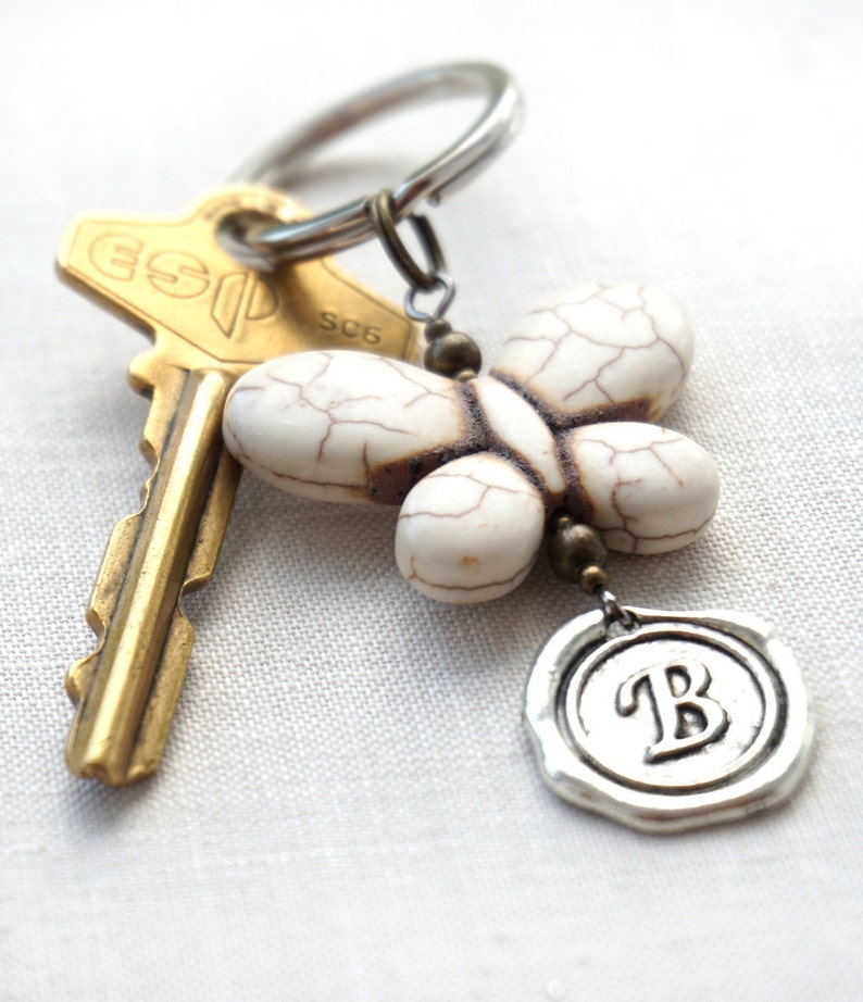 May include: A white butterfly charm with a brown and silver key chain. The charm has a silver seal with the letter "B" on it.