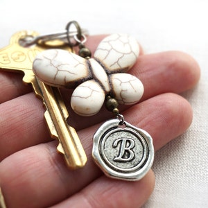 May include: A white butterfly charm with a textured surface hangs from a key ring with a gold key and a silver charm with the letter "B" on it.