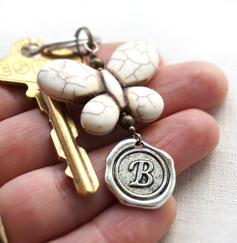 May include: A white butterfly charm with a textured surface hangs from a key ring with a gold key and a silver charm with the letter "B" on it.