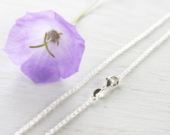 May include: A silver chain necklace with a lobster clasp closure. The chain is delicate and has a fine, rope-like texture. The necklace is photographed on a white wooden surface with a purple flower.