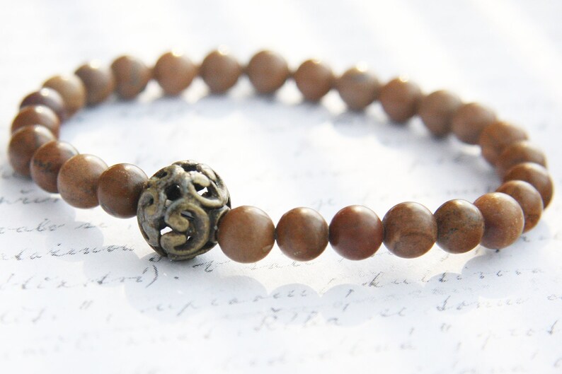 May include: A brown wooden beaded bracelet with a decorative metal charm. The bracelet is made of natural wood beads and features a unique metal charm with a swirling design.