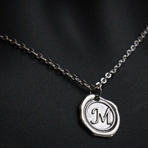 May include: Silver-toned necklace featuring a pendant with the letter "M" inside a circular frame. The pendant has a textured, antique appearance, and hangs from a delicate chain. The necklace is displayed against a dark background.